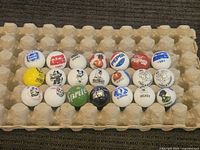 Photo showing 22 white and yellow shooter marbles with colorful vintage advertising logos and cartoon characters laid out on carton holder