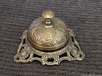 Top and front view of vintage brass dinner bell with floral and geometric embossed patterns on bell dome and intricately designed base with open scrollwork.