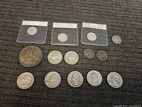 Overview of 15 silver coins including Mercury dimes in plastic holders, several Washington quarters, a large coin, and other smaller coins on a dark fabric surface.