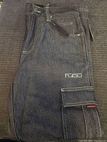 Side view of dark denim FUBU pants showing waistband, front and flap side pocket with logo embroidery.