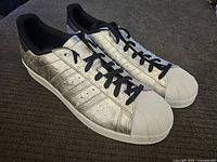 Side angle view showing metallic silver upper and black stripes with black laces; white shell toe and sole visible.