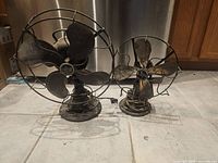 Full view of two antique metal fans side by side on floor, showing overall condition and size difference.