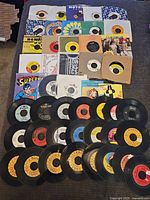Top-down image showing approximately 30 vintage 45 RPM vinyl records laid out on a surface with assorted sleeves and labels visible. Various record labels and colorful sleeves identifiable including Motown, MGM, and other vintage brands.