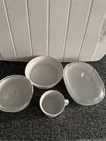 Four white stoneware kitchen pieces: oversized mug, round casserole with lid, oval casserole with lid, and fluted casserole without lid.