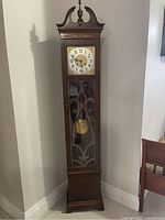 Full front view of the standing clock showing its height, decorative wooden top and glass door with wooden lattice, pendulum visible inside.