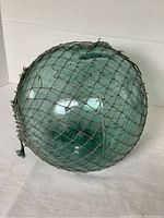 Green glass fishing float ball shown from an angle, featuring netting and tied closure knots.