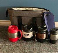 Wide view of all lot items arranged on carpet, showing cooler bag, five insulated mugs and bottle tote in a group