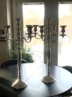 Two large silver metal 5-candle candelabras standing on a black table in front of a window, showing their full height and classic design with five candle holders each.