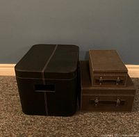 Three storage boxes arranged on carpet with blue wall background. Two fabric covered with handles stacked, smaller box fits inside larger. One black leather-look box with lid and open handles.
