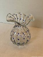 Clear blown art glass vase with blue polka dots and ruffled top edge shown from a side angle on a beige surface.
