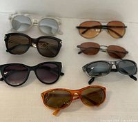 Seven assorted sunglasses displayed on a light surface showing different frame colors, lens tints, and styles.