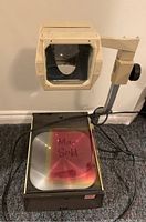 Full view of vintage overhead projector with glass projection surface and transparency sheets.