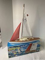 Photo of vintage yacht model with white and red sails standing on original box that acts as display base. Shows wood-look deck and fabric sails.