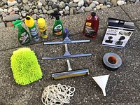 All car cleaning supplies including sprays, squeegees, microfiber mitt, rope, funnel, and duct tape arranged on ground near stones.