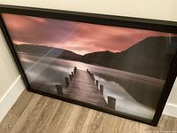 Angle view of the large framed photo leaning against a wall, showing the pier leading into a calm lake with mountains and a colorful sunset in the background. The wooden frame is visible with some glare on the glass.