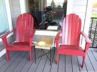 Two red Adirondack chairs and a wicker topped iron side table on a porch deck.
