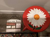 Photo showing red stool with painted white daisy flower and wooden Kitchen Rules sign.