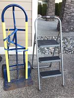 Heavy duty hand truck with a blue metal frame, yellow side guards, blue straps, and two wheels. Next to it is a Rubbermaid aluminum frame step stool with three black textured steps.