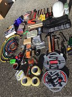 Overview photo of various hand tools, tapes, bungee cords, gloves, safety helmet, and small boxed accessories spread on ground.