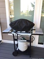 Side view of Weber barbecue covered with black fabric cover on wheeled cart with propane tank.
