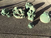 Photo of all seven frog and toad statues arranged outdoors on concrete showing weathered green surfaces and varying sizes and poses.
