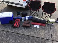 Full view of all camping gear items arranged on pavement including cooler, chairs, backpacks, lantern, tent bag, and boxed item