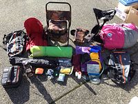 Full lot array showing backpacks, chairs, sleeping bags, camping stove, and accessories spread on ground in sunlight.