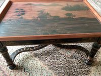 Full view showing the artistic scene under glass on coffee table top, black legs with gold decoration, and patterned carpet under table.