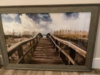 Front view of framed artwork depicting wooden pier leading to beach and ocean with dune grasses and clouds