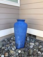 Side view of tall blue ceramic pot placed outdoors among river rocks beside a house.