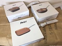 Three white Parini boxes showing the non-stick pans inside. Two round pans with lids and one square griddle pan.