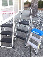 Three step stools and ladder shown outside on concrete, two black Cosco stools with three steps, and one larger aluminum ladder with black steps and gray frame