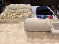 Photo of white king size down comforter with satin edges, folded next to wool blankets and fleece throws in dark colors, plus pillows in packaging and white sheets.