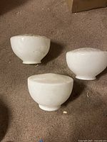Three white milk glass lamp shades placed on carpeted floor showing rounded shapes and base fitting openings.