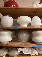 Eight milk glass schoolhouse and globe lampshades on wooden shelves showing different sizes and shapes, some with visible $25 price tags.