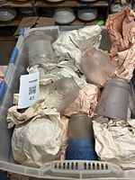Photo of a plastic bin holding multiple antique glass lampshades wrapped in paper. Shades vary in color and finish including frosted, etched, and ribbed glass.