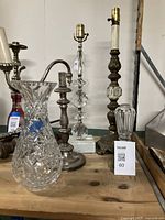 Photo showing five items on a wooden surface: three table lamps with glass and metal components, one cut glass vase, and one silverplate lamp base.