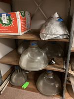 Six Holophane glass lamp shades of varying sizes and shapes on metal shelving, showing ribbed and prismatic glass design typical for industrial lighting.