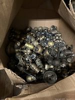 Open cardboard box filled with brass and metal antique lamp socket clusters and parts including pull chains and socket housings.
