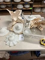 View of multiple milk glass smoke bells of various fluted and ruffled designs, some wrapped in packing paper on table.