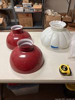 Three oil lamp glass shades on a table: two red student lamp shades and one white milk glass shade.
