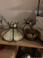 Two stained glass panel lamps and part of a third on a wooden shelf with metal frame, showing their cream and amber tinted glass panels with metal beading and fringed bases.