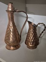 Two small copper jugs, one taller with hinged lid and geometric pattern, one smaller with scale texture and twisted handle