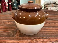 Front and side view of the glossy brown and off-white ceramic bean pot with lid on wooden surface.