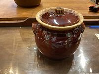 Brown ceramic bean pot with lid placed on a wooden surface reflecting light.