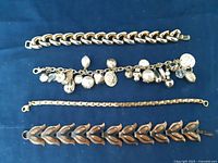 Four bracelets laid out on blue fabric: gold chunky curb chain, charm bracelet with dangling charms, copper link bracelet with leaf designs, tri-color chain bracelet