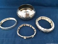 Four silver tone bracelets of different styles arranged on a dark fabric background. Visible are a wide rounded bangle with engraved design, a thin smooth oval bangle, a rectangular link bracelet, and a thick patterned link bracelet.