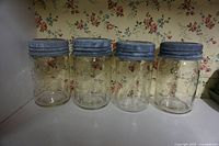 Front view of four vintage Crown glass mason jars with blue metal screw band lids lined up in a row on a white shelf against floral wallpaper.