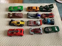 Top view of 12 Hot Wheels cars arranged in four rows of three on a beige mat