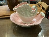 Photo of large pink clam shell shaped ceramic basin and matching iridescent shell-shaped jug with gold handle on table.
